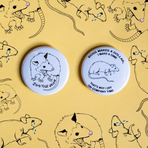 May include: Two round white buttons with black text and illustrations. The button on the left features a black and white illustration of an opossum with the text "Join Your Union". The button on the right features a black and white illustration of a rat with the text "Boss Makes a Dollar, I Make a Dime. That's Why I Cry On Company Time."