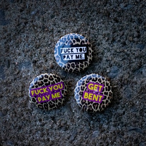 May include: Three round buttons with leopard print backgrounds and bold text. The buttons display phrases like "FUCK YOU PAY ME" and "GET BENT" in white and purple. The buttons are set against a textured, gray surface.