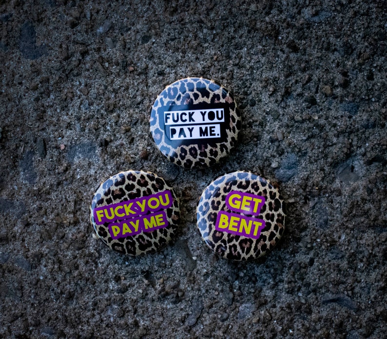 May include: Three round buttons with leopard print backgrounds and bold text. The buttons display phrases like "FUCK YOU PAY ME" and "GET BENT" in white and purple. The buttons are set against a textured, gray surface.