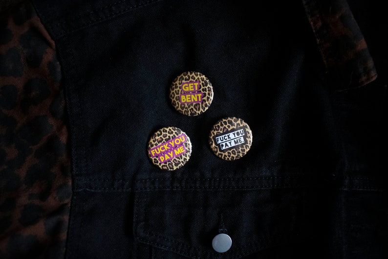 May include: Three round buttons with leopard print backgrounds and bold text. The buttons say "GET BENT", "FUCK YOU PAY ME", and "FUCK YOU PAY ME". The buttons are on a black denim jacket with a leopard print accent.