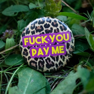 May include: A round button with a leopard print background. The button has the text "FUCK YOU PAY ME" in yellow letters on a purple background. The button is surrounded by green leaves and grass.