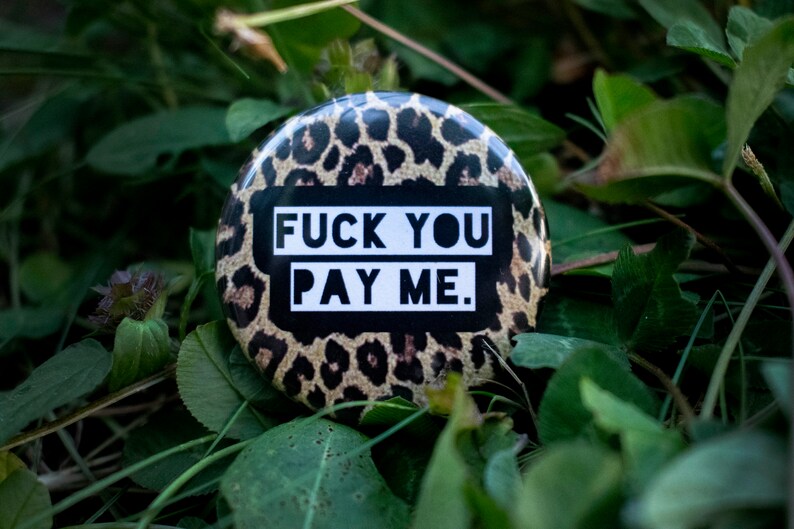 May include: A round button with a leopard print background and the text "FUCK YOU PAY ME" in bold white letters on a black rectangle. The button is set against a backdrop of green foliage.