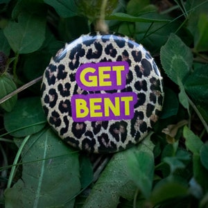 May include: A round button with a leopard print background and the words "GET BENT" in yellow letters on a purple background. The button is set against a backdrop of green leaves and grass.