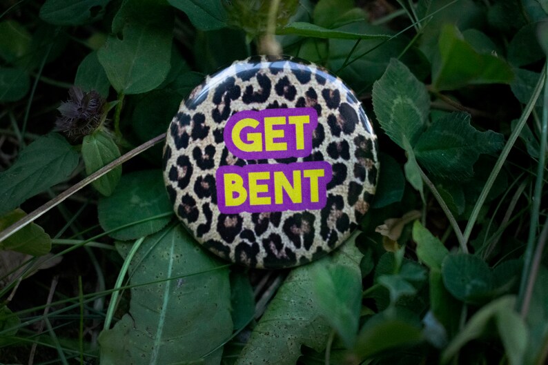 May include: A round button with a leopard print background and the words "GET BENT" in yellow letters on a purple background. The button is set against a backdrop of green leaves and grass.