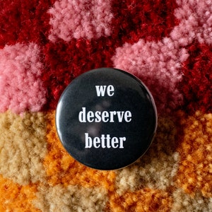 May include: Black button with white text that reads "we deserve better" on a colourful fuzzy background.