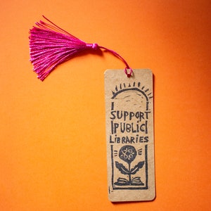Puede incluir: Un marcapáginas marrón con un borla rosa. El marcapáginas tiene una ilustración en blanco y negro de una flor que crece de un libro con el texto "Support Public Libraries".
