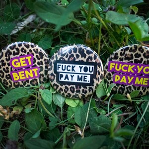 May include: Three round buttons with a leopard print background. Each button has a different phrase in bold, colorful text: "GET BENT", "FUCK YOU PAY ME", and "FUCK YOU PAY ME". The buttons are set among green foliage.