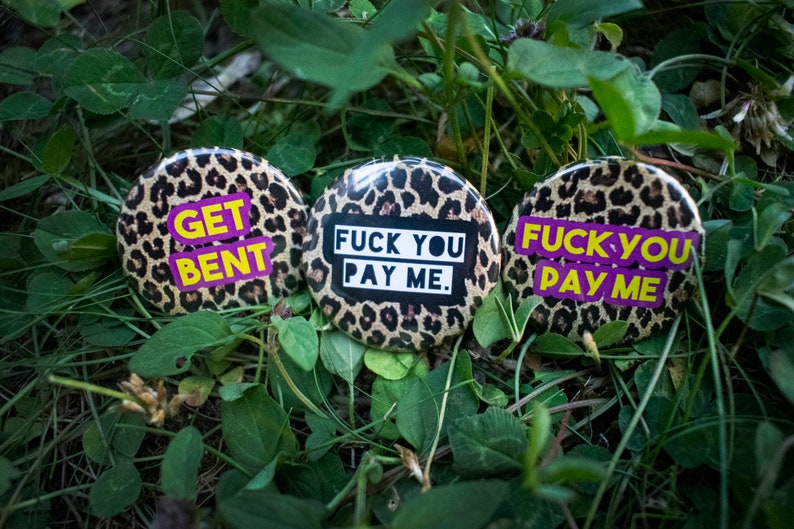 May include: Three round buttons with a leopard print background. Each button has a different phrase in bold, colorful text: "GET BENT", "FUCK YOU PAY ME", and "FUCK YOU PAY ME". The buttons are set among green foliage.