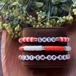May include: Three beaded bracelets with white and orange beads. The top bracelet spells out "SHSUD", the middle bracelet is white and orange, and the bottom bracelet spells out "EATEMUPKAT". The bracelets are displayed on a brown stand.
