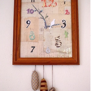 May include: A wooden framed clock with a collage of paper scraps and fabric numbers. The clock has three fabric pendulum weights hanging from it.