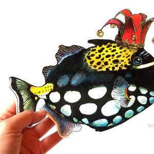 May include: A black, white, and yellow fish with a jester's hat on its head. The fish has a round body with white spots and a long, thin tail. The jester's hat is red and yellow with bells.