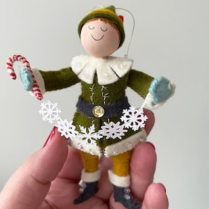 Elf dressed in a green coat and yellow tights holding a string of snowflakes and a candy cane.
