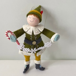 Elf Christmas ornament made of felt. They are dressed in green and yellow with white trim and grey boots. They are holding a candy cane and snowflakes.