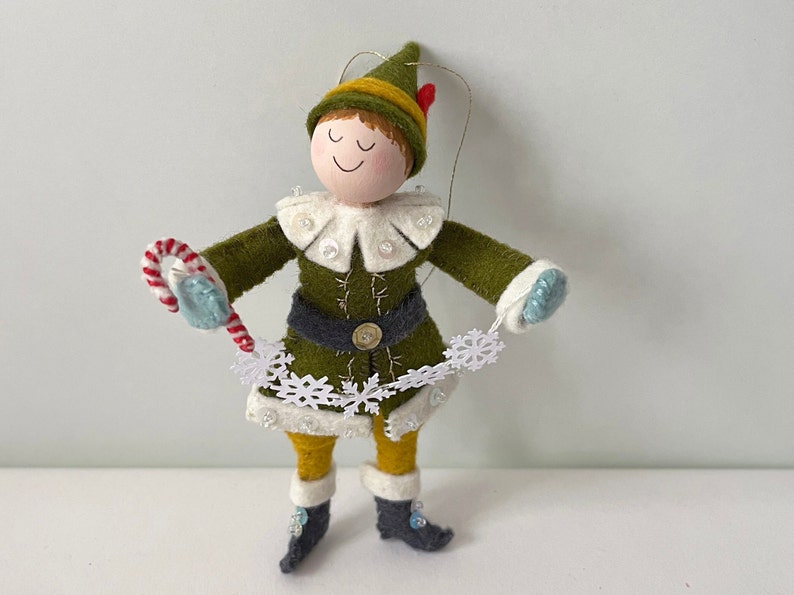 Elf Christmas ornament made of felt. They are dressed in green and yellow with white trim and grey boots. They are holding a candy cane and snowflakes.