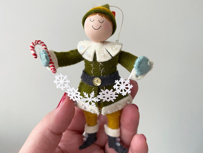 Elf dressed in a green coat and yellow tights holding a string of snowflakes and a candy cane.