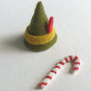 View of tiny elf accessories. A green felt hat with a yellow stripe and red feather. There is also a tiny candy cane made of a pipe cleaner and red thread.