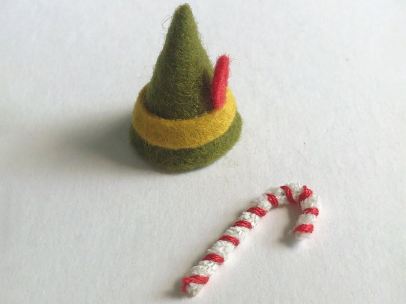 View of tiny elf accessories. A green felt hat with a yellow stripe and red feather. There is also a tiny candy cane made of a pipe cleaner and red thread.