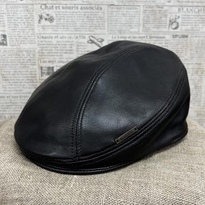 May include: A black leather newsboy cap with a stitched design and a small metal logo that reads "Durbano".