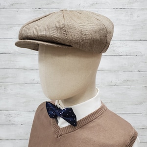 Pode incluir: Um bon&eacute; newsboy marrom de sarja de espinha de peixe com aba ligeiramente curvada. O bon&eacute; est&aacute; em um manequim que est&aacute; vestindo um su&eacute;ter marrom e uma camisa branca com uma gravata borboleta azul marinho.