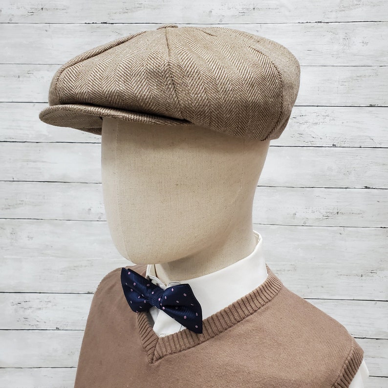 Pode incluir: Um bon&eacute; newsboy marrom de sarja de espinha de peixe com aba ligeiramente curvada. O bon&eacute; est&aacute; em um manequim que est&aacute; vestindo um su&eacute;ter marrom e uma camisa branca com uma gravata borboleta azul marinho.