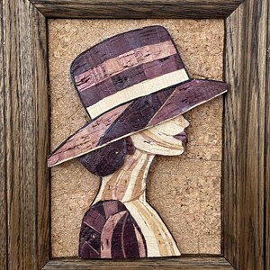 Puede incluir: Una obra de arte de mosaico de corcho enmarcada que representa a una mujer con un sombrero de ala ancha. El sombrero y la cara de la mujer están hechos de piezas de corcho en tonos marrones, beige y morado.