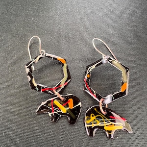 May include: A pair of dangle earrings featuring a black hexagonal frame and a black bear-shaped charm. Both components are decorated with abstract splatters of red, yellow, orange, and white paint. The earrings hang from silver-toned metal hooks.