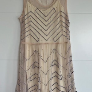 May include: A sleeveless, cream-colored dress with a sheer overlay. The dress features a chevron pattern of dark gray beads across the bodice and skirt. The dress is hanging on a black hanger against a white wall.