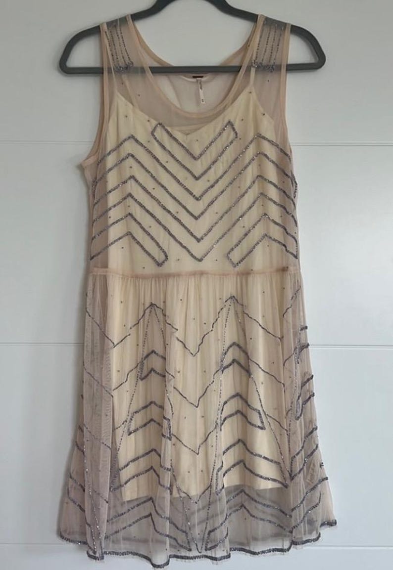 May include: A sleeveless, cream-colored dress with a sheer overlay. The dress features a chevron pattern of dark gray beads across the bodice and skirt. The dress is hanging on a black hanger against a white wall.