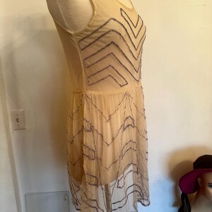 May include: A beige, sleeveless dress with a sheer overlay. The dress features a zigzag pattern of silver beading across the bodice and skirt. The dress is displayed on a mannequin.