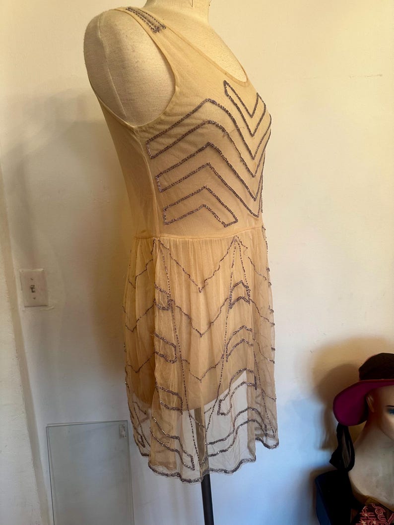 May include: A beige, sleeveless dress with a sheer overlay. The dress features a zigzag pattern of silver beading across the bodice and skirt. The dress is displayed on a mannequin.