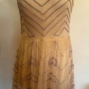 May include: A beige, sleeveless dress with a layered skirt. The dress is adorned with silver-colored beadwork in a geometric pattern across the bodice and skirt. The dress is displayed on a mannequin.