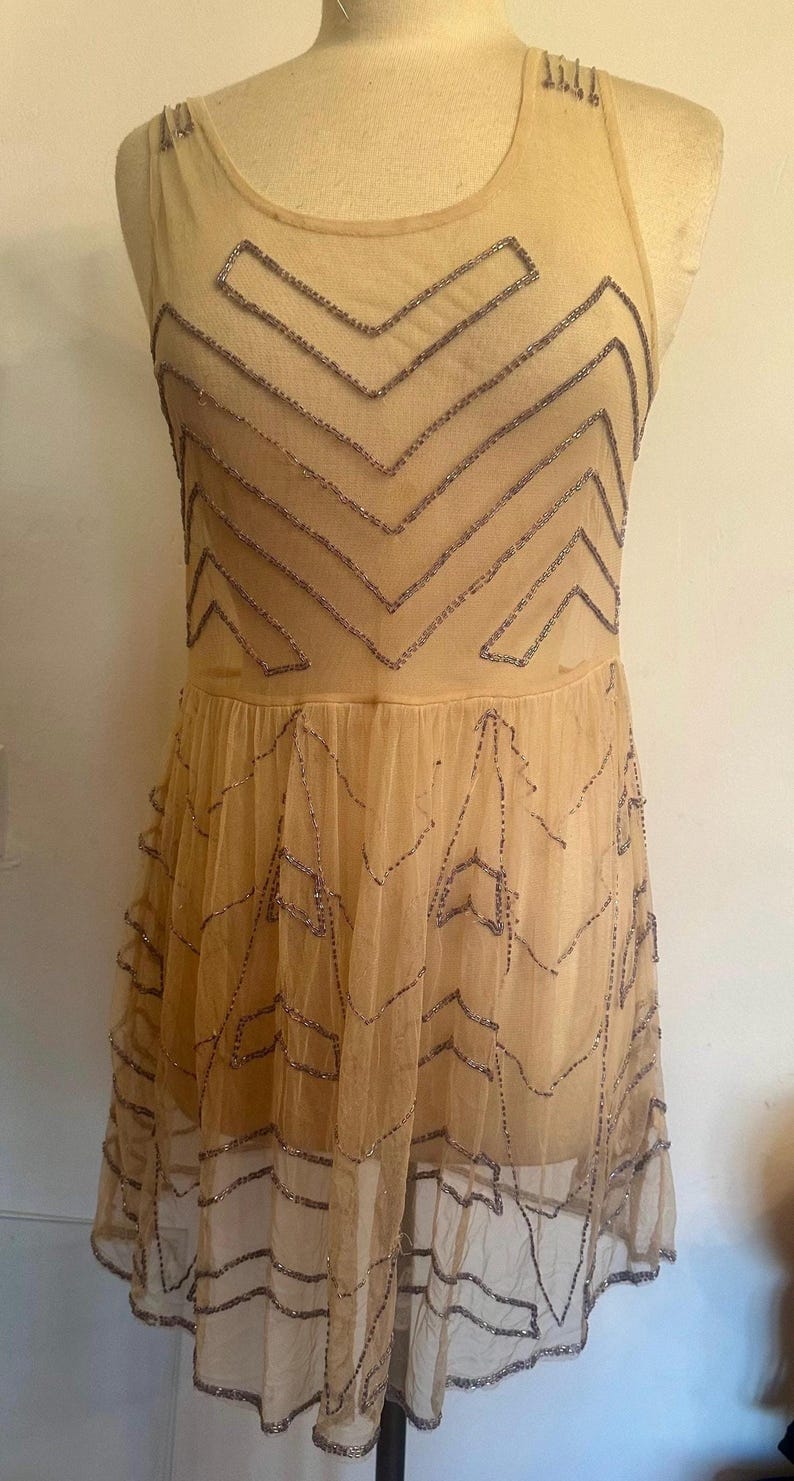 May include: A beige, sleeveless dress with a layered skirt. The dress is adorned with silver-colored beadwork in a geometric pattern across the bodice and skirt. The dress is displayed on a mannequin.