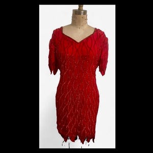 May include: A red, short-sleeved dress with a V-neckline. The dress is adorned with a pattern of vertical, beaded lines. The hemline has a scalloped edge. The dress is displayed on a mannequin.