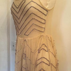 May include: A vintage-style, beige dress with a sheer overlay, adorned with intricate beaded patterns in a chevron design. The dress is sleeveless and appears to be made of delicate fabric, possibly silk or chiffon. The dress is displayed on a mannequin.