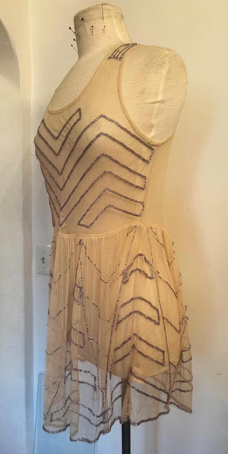 May include: A vintage-style, beige dress with a sheer overlay, adorned with intricate beaded patterns in a chevron design. The dress is sleeveless and appears to be made of delicate fabric, possibly silk or chiffon. The dress is displayed on a mannequin.