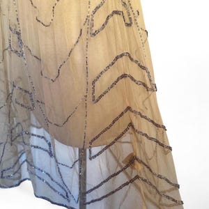 May include: A sheer, beige fabric skirt with intricate, geometric patterns created by rows of silver beads. The skirt appears to be layered, with the beaded design adding texture and visual interest. The skirt is likely vintage or inspired by vintage designs.