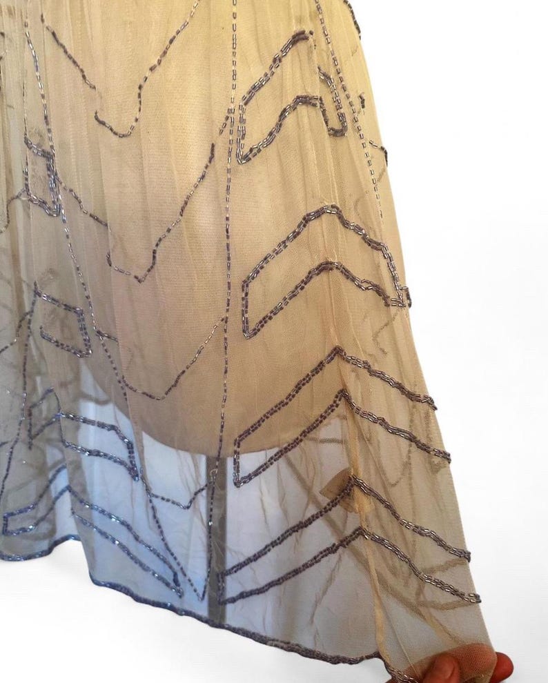 May include: A sheer, beige fabric skirt with intricate, geometric patterns created by rows of silver beads. The skirt appears to be layered, with the beaded design adding texture and visual interest. The skirt is likely vintage or inspired by vintage designs.