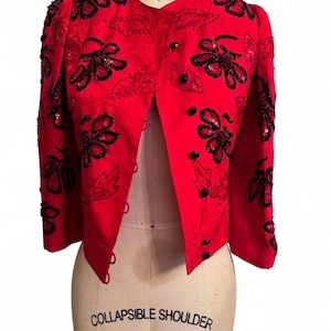 Può includere: Giacca rossa con decorazioni floreali nere e bottoni neri. La giacca è esposta su un manichino. Ha uno scollo a V e maniche a tre quarti. Decorata con paillettes e perline nere.
