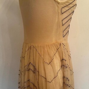 May include: A vintage-style, beige dress on a mannequin. The dress features a fitted bodice and a flowing skirt. The dress is adorned with beaded embellishments in a zigzag pattern, adding a touch of glamour to the overall design.