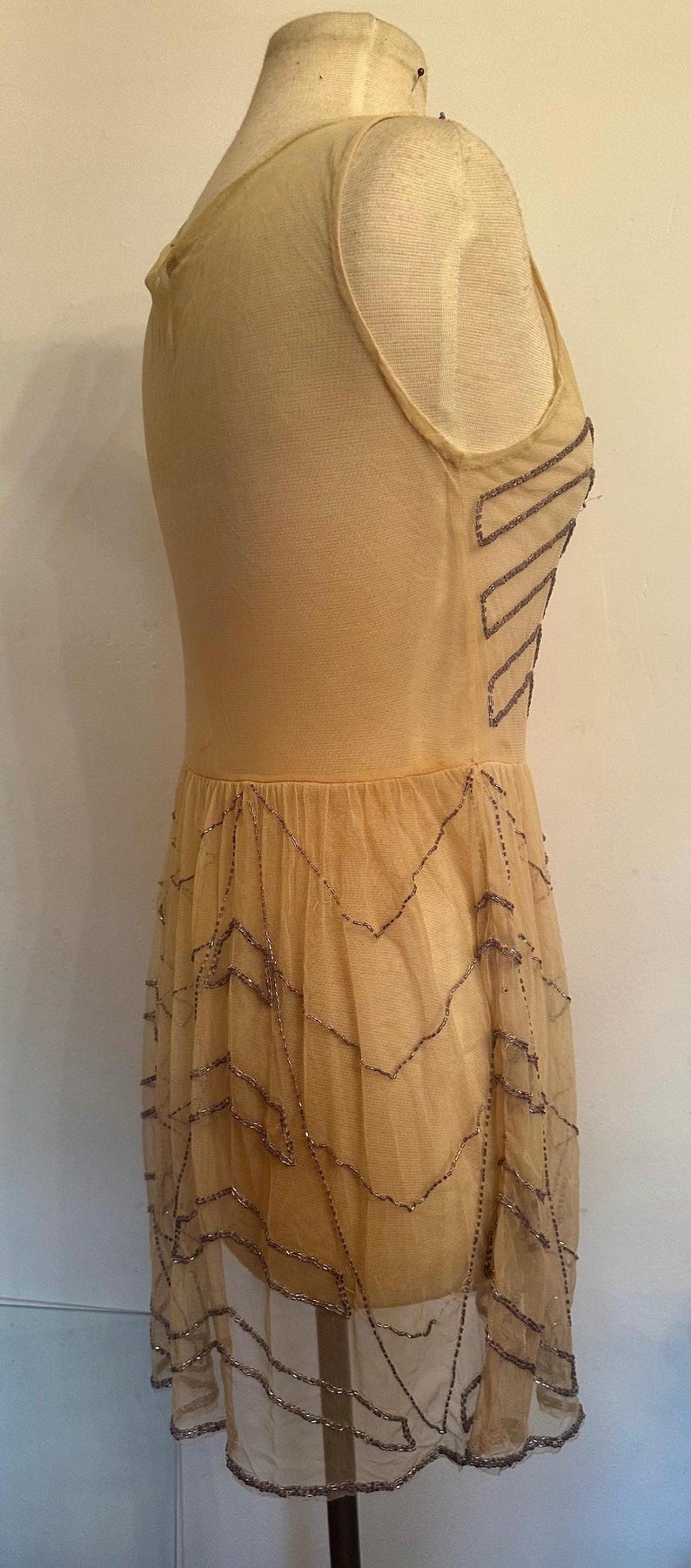 May include: A vintage-style, beige dress on a mannequin. The dress features a fitted bodice and a flowing skirt. The dress is adorned with beaded embellishments in a zigzag pattern, adding a touch of glamour to the overall design.