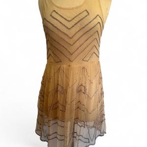May include: A tan-colored, sleeveless dress with a sheer overlay. The dress features a geometric pattern of dark beads across the bodice and skirt. The dress is displayed on a mannequin.
