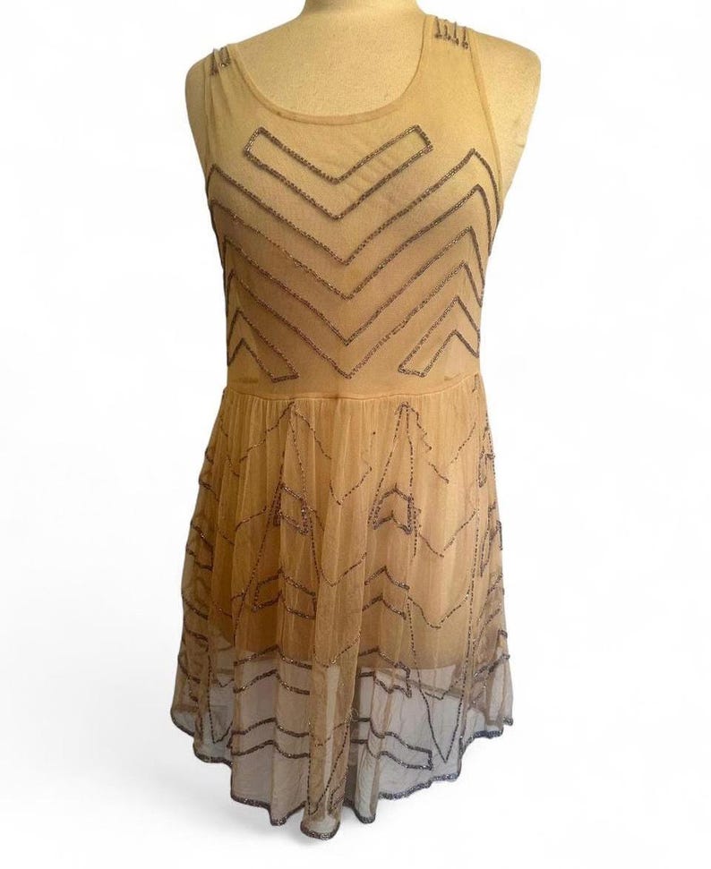 May include: A tan-colored, sleeveless dress with a sheer overlay. The dress features a geometric pattern of dark beads across the bodice and skirt. The dress is displayed on a mannequin.