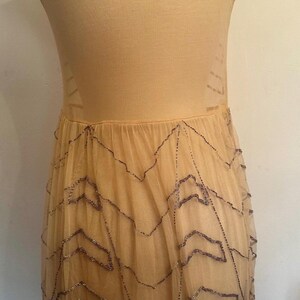 May include: A beige sleeveless dress on a mannequin. The dress features a fitted bodice and a flowing skirt made of sheer fabric. The skirt is adorned with a beaded zigzag pattern. The dress is a neutral color.