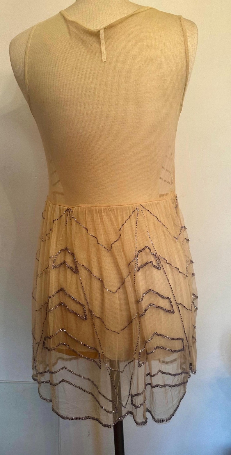 May include: A beige sleeveless dress on a mannequin. The dress features a fitted bodice and a flowing skirt made of sheer fabric. The skirt is adorned with a beaded zigzag pattern. The dress is a neutral color.