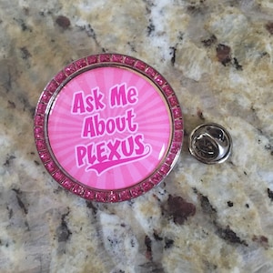 May include: Pink and silver round pin with rhinestones around the edge. The pin has a pink background with white text that says "Ask Me About Plexus".
