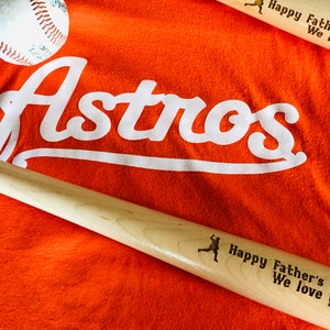 May include: Orange baseball t-shirt with the word "Astros" in white lettering. Two wooden baseball bats with the text "Happy Father's Day #1 Dad We love you!" are on top of the shirt.