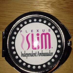 May include: A silver compact mirror with a black and white design. The mirror has a white background with the text "PLEXUS Slim. Independent Ambassador." in black and pink lettering. The text is surrounded by a black circle with white dots.