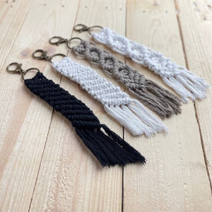 Op de afbeelding: Vier macramé sleutelhangers met kwastjes in zwart, wit en beige. De sleutelhangers zijn gemaakt met een knooptechniek en hebben een metalen ring.