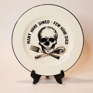 May include: A white ceramic dinner plate with a black rim and a black and white skull and crossbones design. The plate has the text "Many have dined. Few have died." around the skull.