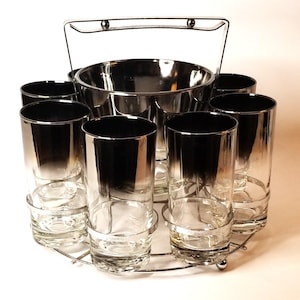 May include: A set of eight silver-rimmed glasses with a black ombre effect, a silver ice bucket with a handle, and a silver metal stand. The glasses are arranged in a circle around the ice bucket.